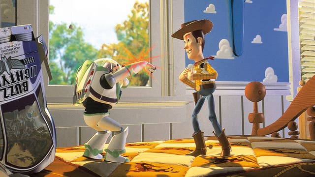 Eine animierte Szene zeigt zwei Spielzeugfiguren in einem Kinderzimmer. Links steht eine Figur in einem Raumanzug, die mit einem ausgestreckten Arm einen roten Laserstrahl abfeuert. Rechts steht eine Cowboy-Figur mit einem Hut, die lächelnd zuschaut. Im Hintergrund ist ein Fenster mit Blick auf Bäume und ein blauer Himmel mit Wolkenmuster an der Wand zu sehen. Auf dem Bett liegt eine bunte Decke mit Cowboy-Motiven. Links im Bild ist eine Verpackung mit der Aufschrift "Buzz Lightyear" zu erkennen.