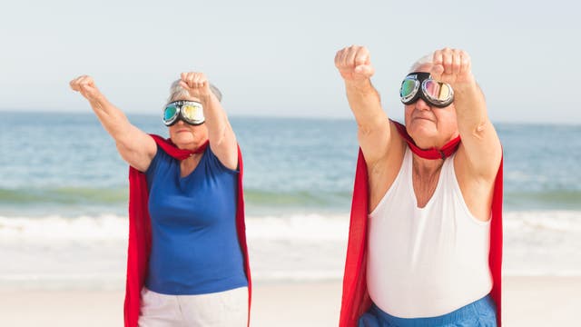 Zwei ältere Personen stehen am Strand und posieren mit ausgestreckten Armen, als ob sie fliegen würden. Sie tragen rote Umhänge und Schutzbrillen, was an Superhelden erinnert. Im Hintergrund ist das Meer zu sehen.