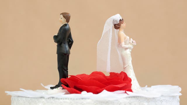 Auf einer Hochzeitstorte stehen zwei Plastikfiguren von Braut und Bräutigam, jedoch mit Abstand und einander den Rücken zugewandt, dazwischen eine rote Rose.