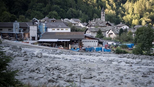 Eine kleine, von Bäumen umgebene Ortschaft mit traditionellen Steinhäusern und einer Kirche im Hintergrund. Im Vordergrund sind Industriegebäude und Container zu sehen. Der Boden ist mit Geröll und Schlamm bedeckt, was auf einen Erdrutsch oder eine Überschwemmung hindeutet. 