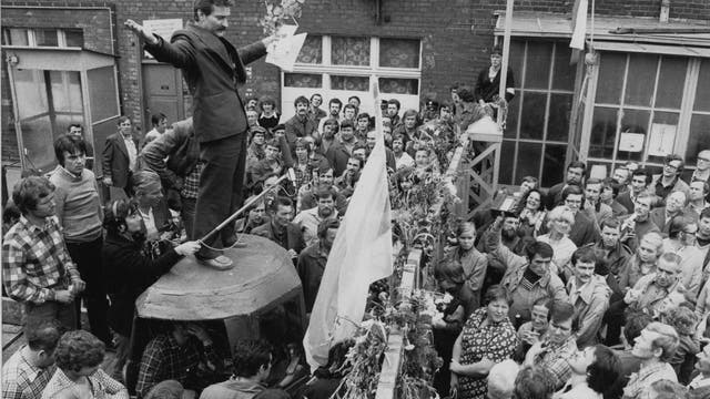 Der junge Lech Wałęsa steht auf einem kleinen Podest inmitten einer Menge von Männern und Frauen. Er trägt ein dunkles Sakko und hat beide Arme seitlich vom Körper weggestreckt.