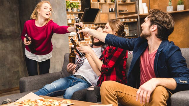 Eine Gruppe von vier jungen Erwachsenen in einem gemütlichen Wohnzimmer mit Backsteinwänden und Pflanzen im Hintergrund. Eine Frau steht und reicht Bierflaschen, die drei anderen sitzen auf einer Couch. Auf dem Tisch vor ihnen liegen zwei Pizzen. Die Szene vermittelt eine entspannte und fröhliche Atmosphäre.