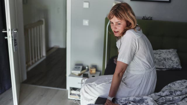 Eine Person in einem weißen Kleid sitzt auf einem Bett in einem Schlafzimmer. Der Raum ist in neutralen Tönen gehalten, mit einem Nachttisch, auf dem Bücher und kleine Gegenstände liegen. Eine offene Tür führt zu einem Flur. Die Person schaut nachdenklich zur Seite. Im Hintergrund hängt ein gerahmtes Bild an der Wand.