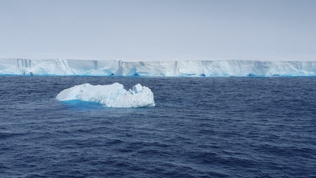 Eine weiße Eiswand ragt aus dem blauen Südpolarmeer, in dem ein viel kleinerer Eisberg zusätzlich schwimmt.