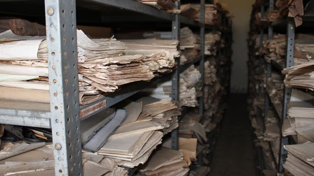 Regale voller alter Papierdokumente in einem Archivraum in der Demokratischen Republik Kongo. Die Dokumente sind gestapelt und teilweise vergilbt. Im Raum stehen mehrere Reihen von Metallregalen eng nebeneinander.