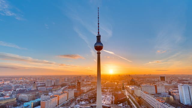Stadtansicht von Berlin bei Sonnenuntergang, mit dem Fernsehturm im Vordergrund. Der Himmel ist klar mit vereinzelten Wolken, und die Sonne geht am Horizont unter, was die Gebäude in warmes Licht taucht. Die Stadtlandschaft zeigt eine Mischung aus modernen und historischen Gebäuden.