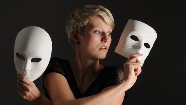 Eine Frau hält zwei unterschiedliche weiße Masken rechts und links von sich vor schwarzem Hintergrund.