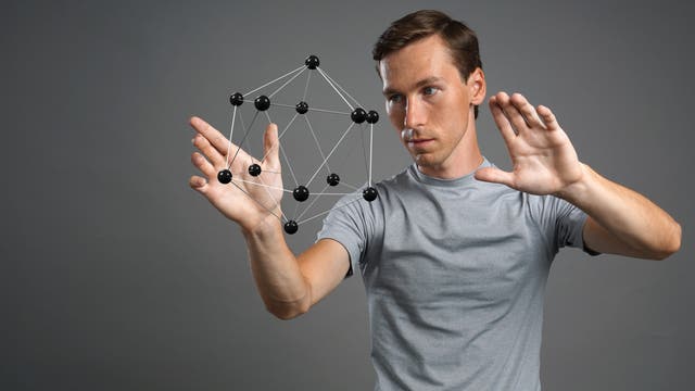 Ein Mann im grauen T-Shirt steht neben einem molekülähnlichen Dodekaeder aus Kugeln und Stäben.