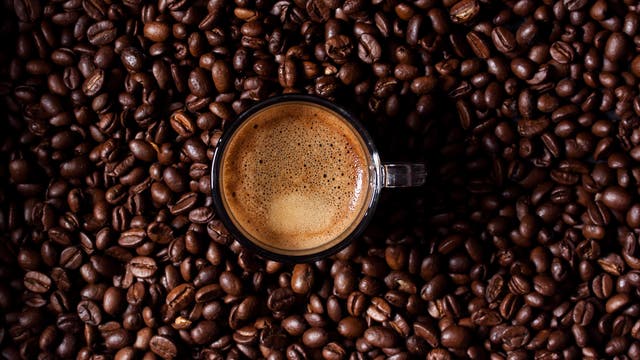 Eine Tasse Espresso mit einer cremigen Schaumkrone steht auf einer Oberfläche, die vollständig mit gerösteten Kaffeebohnen bedeckt ist.