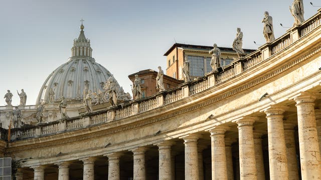 Eine Ansicht der Kolonnaden des Petersplatzes im Vatikan, die von zahlreichen Statuen gesäumt sind. Im Hintergrund ist die Kuppel des Petersdoms sichtbar. Die Architektur zeigt klassische Säulen und barocke Details. Der Himmel ist klar und blau. Keine Personen sind im Bild zu sehen.