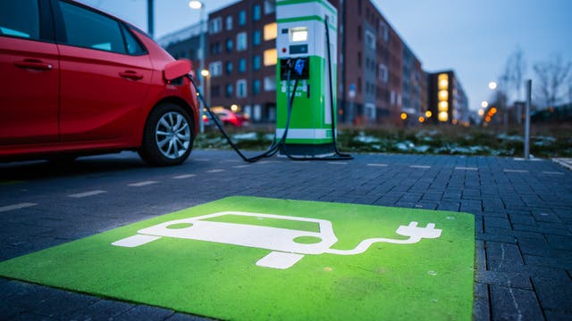 Ein rotes Elektroauto ist an eine grüne Ladestation angeschlossen, die sich auf einem Parkplatz in einer städtischen Umgebung befindet. Der Boden ist mit einem grünen Symbol für Elektrofahrzeuge markiert. Im Hintergrund sind moderne Wohngebäude und Straßenlaternen zu sehen. Die Szene vermittelt eine nachhaltige und umweltfreundliche Mobilität.