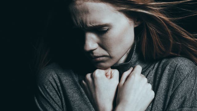 Eine Frau in einem dunklen Raum hält ihre Hände vor der Brust und blickt nach unten. Der Gesichtsausdruck wirkt nachdenklich oder besorgt. 
