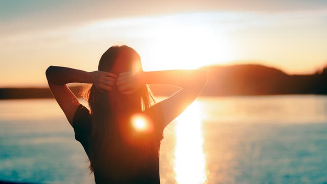Man sieht eine Frau mit verschränkten Armen von hinten, sie genießt den Sonnenuntergang am See.