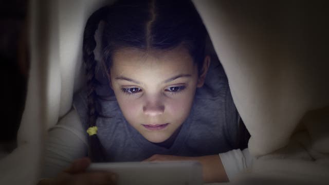Ein junges Mädchen liegt unter einer Bettdecke und guckt konzentriert auf ein Bildschirmgerät.