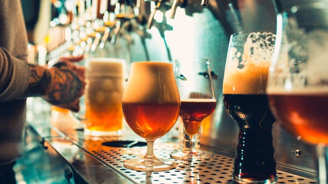 Eine Reihe von Biergläsern mit verschiedenen Biersorten steht auf einer Theke in einer Bar. Im Hintergrund ist eine Person zu sehen, die ein weiteres Glas unter einen Zapfhahn hält. Die Gläser sind unterschiedlich geformt und gefüllt, einige mit Schaumkronen. Die Szene ist in warmem Licht gehalten, das die Farben der Biere betont.