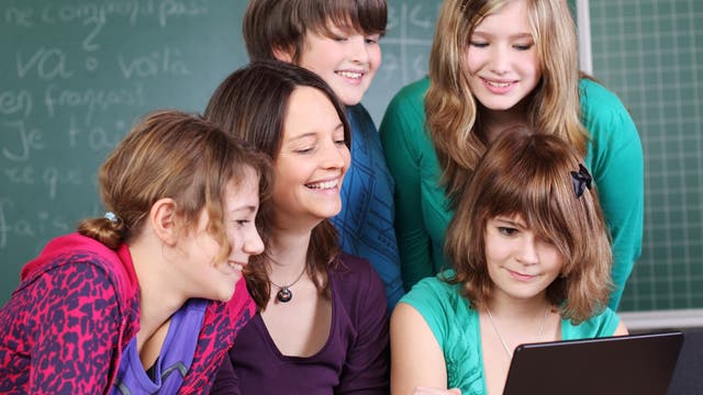 Eine Lehrerin und 4 Schüler*innen schauen vergnügt lächelnd in einen Laptop.