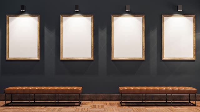 Eine Galerie mit vier leeren, beleuchteten Bilderrahmen an einer dunkelgrauen Wand. Darunter befinden sich zwei braune, gepolsterte Bänke auf einem Parkettboden. 