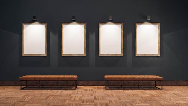 Vier leere, beleuchtete Bilderrahmen hängen an einer dunkelgrauen Wand in einer Galerie. Darunter stehen zwei braune Bänke auf einem Parkettboden. 