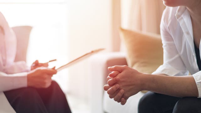 Zwei Personen sitzen in einem hellen Raum. Die Person links hält einen Stift und ein Klemmbrett, während die Person rechts die Hände gefaltet hat. Beide tragen weiße Blusen. Die Szene deutet auf ein Gespräch oder eine Beratung hin. Im Hintergrund sind verschwommen Möbel zu sehen.