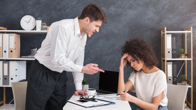Ein Mann in einem weißen Hemd steht in einem Büro und zeigt mit dem Finger auf eine Frau, die mit besorgtem Gesichtsausdruck am Schreibtisch sitzt. Vor ihr stehen ein Laptop, eine Tasse Kaffee und Dokumente. Im Hintergrund sind Regale mit Ordnern und eine Uhr an der Wand zu sehen. Die Szene vermittelt eine angespannte Arbeitsatmosphäre.