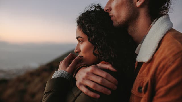 Ein Paar schaut in der Natur Richtung Horizont. Der Mann hält seine Partnerin fest in den Armen.
