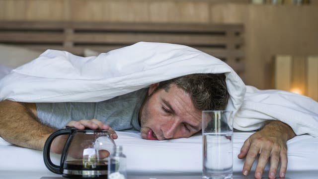 Ein verkaterter Mann guckt unter einer Bettdecke hervor.
