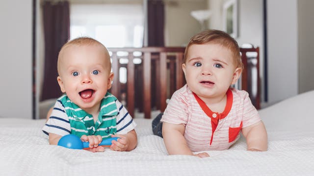 Zwei Babys liegen auf einem Bett. Das Baby links trägt ein gestreiftes Shirt, lächelt und hält ein blaues Spielzeug. Das Baby rechts trägt ein gestreiftes Shirt mit rotem Kragen und sieht besorgt aus. Im Hintergrund sind ein Kinderbett und ein Fenster zu sehen.