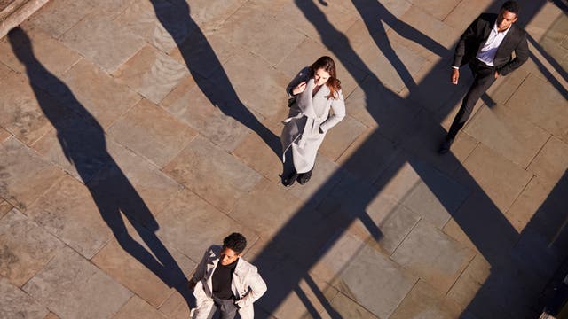 Drei Personen gehen auf einem urban wirkend gepflasterten Weg, ihre langen Schatten fallen auf den Boden. Eine Person in einem grauen Mantel hält ein Handy ans Ohr, während die anderen beiden in Anzügen zügig gehen. Alle 3 Personen wirken wie Geschäftsleute. Die Szene wird von oben betrachtet, was die Schatten betont.
