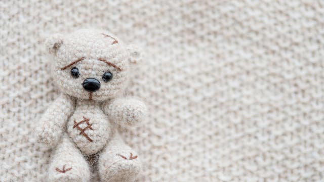 Liegender kleiner gestrickter wollweißer Babybär mit traurigen Knopfaugen auf einer weißen Wolldecke.