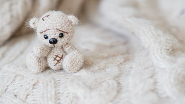 Kleiner gestrickter wollweißer Babybär mit traurigen Knopfaugen sitzt auf einer weißen Strickdecke.