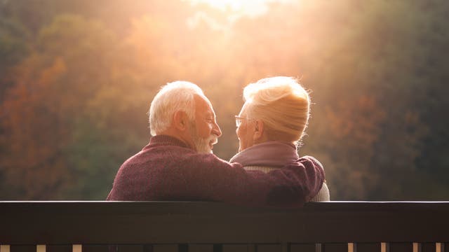 Ein älteres Paar sitzt auf einer Bank und schaut sich liebevoll an. Die Szene ist in warmes, goldenes Licht getaucht, das von der tiefstehenden Sonne im Hintergrund erzeugt wird. Im Hintergrund sind unscharfe Herbstbäume zu sehen, die eine ruhige und friedliche Atmosphäre schaffen.