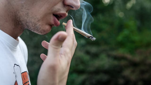 Eine Person hält einen rauchenden Joint nahe am Mund. Der Hintergrund ist unscharf und zeigt grüne Vegetation. Die Person trägt ein weißes T-Shirt mit einem teilweise sichtbaren Aufdruck. Rauch steigt vom Joint auf.