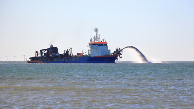 Ein großes blaues Schiff, das als Saugbagger fungiert, befindet sich auf dem Meer. Es sprüht Wasser und Sand in einem Bogen zurück ins Meer. Im Hintergrund sind Windkraftanlagen am Horizont zu sehen. Der Himmel ist klar und blau.