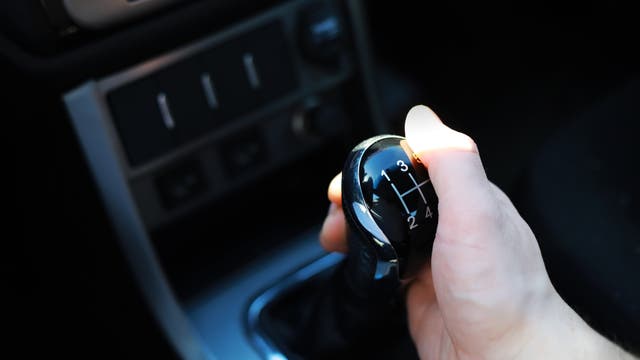 Eine Hand hält den Schalthebel eines Autos, der die Gänge 1 bis 5 und den Rückwärtsgang anzeigt. Der Schalthebel befindet sich in einem Fahrzeugcockpit mit sichtbaren Bedienelementen im Hintergrund.