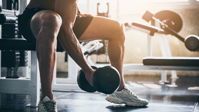 Ein Mann hebt im Fitnessstudio ein Gewicht an. Die Sonne scheint auf seinen muskulösen Oberarm 