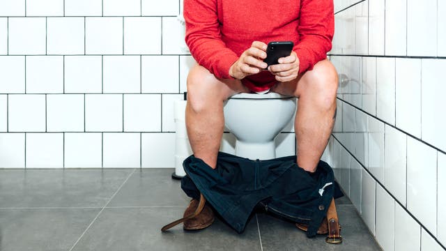 Eine Person in einem roten Pullover sitzt auf einer Toilette in einem gefliesten Badezimmer und hält ein Smartphone in den Händen. Die Hose ist heruntergelassen, und die Person scheint auf das Telefon zu schauen.