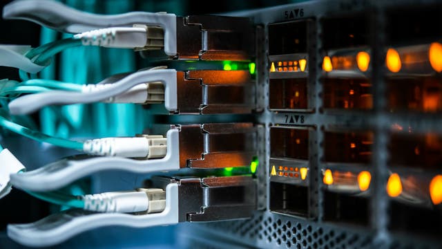 Nahaufnahme eines Serverracks mit mehreren eingesteckten Netzwerkkabeln. Die Kabel sind mit leuchtenden grünen und orangefarbenen LED-Anzeigen verbunden, die den Netzwerkstatus anzeigen. Die Umgebung ist in einem gedämpften, bläulichen Licht gehalten, das die technische Atmosphäre unterstreicht.