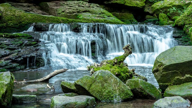 Ein kleiner Wasserfall fließt über moosbedeckte Felsen in einem grünen, bewaldeten Gebiet. Im Vordergrund liegen große Steine und ein mit Moos bewachsener Baumstumpf im Wasser. Die Umgebung ist üppig und grün.