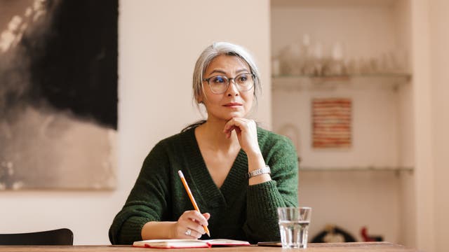 Eine grauhaarige Frau sitzt am Schreibtisch und denkt nach, während sie sich Notizen macht