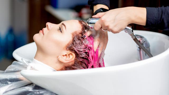 Eine Person liegt in einem Friseursalon an einem Waschbecken, während eine andere Person ihr die Haare wäscht. Die Haare sind mit einer pinken Farbe behandelt. Die Szene zeigt den Prozess des Haarewaschens mit einem Duschkopf. Im Hintergrund sind unscharfe Details des Salons zu sehen.