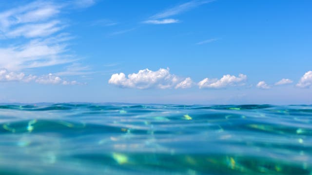 Eine ruhige Meeresoberfläche mit sanften Wellen im Vordergrund, die das Licht reflektieren. Im Hintergrund erstreckt sich ein klarer blauer Himmel mit vereinzelten weißen Wolken. Die Szene vermittelt ein Gefühl von Ruhe und Weite.