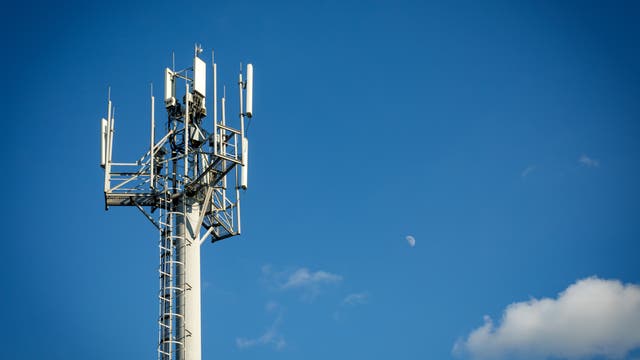 Ein Mobilfunkmast mit mehreren Antennen ragt in den klaren blauen Himmel. Im Hintergrund ist der Mond sichtbar, umgeben von wenigen kleinen Wolken. Der Mast symbolisiert moderne Kommunikationstechnologie.