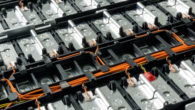 Nahaufnahme einer Batterieeinheit mit mehreren Zellen, verbunden durch orangefarbene Kabel und Metallverbindungen. Die Struktur zeigt eine Anordnung von rechteckigen Modulen, die in einem Rastermuster angeordnet sind. Die Oberfläche der Zellen ist metallisch glänzend, und die Kabel sind sorgfältig in schwarzen Halterungen fixiert. Keine sichtbaren Texte oder URLs.