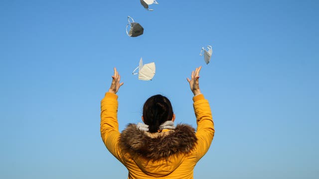 Eine Frau von hinten in gelber Winterjacke, die vor einem blauen Himmel mehrere FFP2-Masken in die Luft wirft.