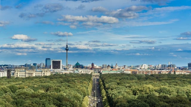Blick auf die Skyline von Berlin mit dem markanten Fernsehturm und dem Brandenburger Tor im Zentrum. Im Vordergrund erstreckt sich der Tiergarten, ein großer, grüner Park, durch den eine Straße mit fahrenden Autos führt. Die Stadtlandschaft dahinter zeigt eine Mischung aus historischen und modernen Gebäuden. Der Himmel ist blau mit vereinzelten Wolken.