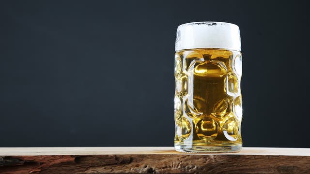 Ein großes, mit goldenem Bier gefülltes Glas mit Schaumkrone steht auf einem rustikalen Holztisch vor einem dunklen Hintergrund. Das Glas hat ein markantes, strukturiertes Design, das das Licht reflektiert.
