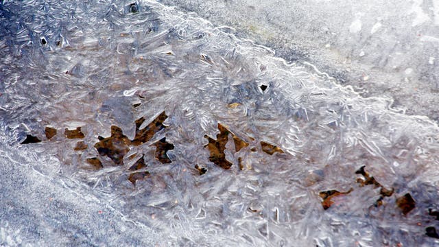 Eine Nahaufnahme von gefrorenem Eis auf einer Oberfläche, das komplexe, kristalline Muster bildet. Die Eisstruktur zeigt verschiedene Formen und Texturen, die durchscheinend und schimmernd wirken. Darunter sind dunklere Bereiche sichtbar, die möglicherweise Erde oder Wasser darstellen. Die Szene vermittelt ein Gefühl von Kälte und natürlicher Schönheit.