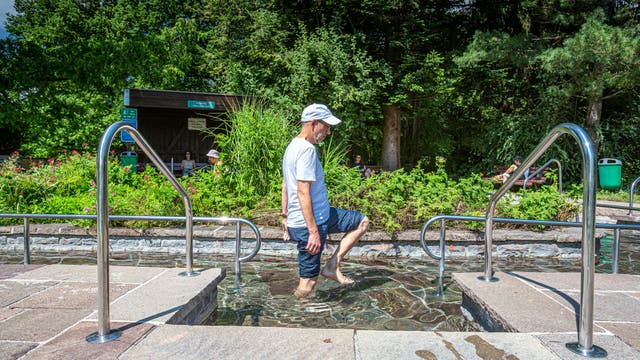 Eine Person geht durch ein Kneippbecken im Freien, umgeben von üppigem Grün und Bäumen. Der Himmel ist klar, und die Sonne scheint. Im Hintergrund sind weitere Menschen auf Bänken zu sehen. Edelstahlgeländer flankieren den Weg durch das Wasser.