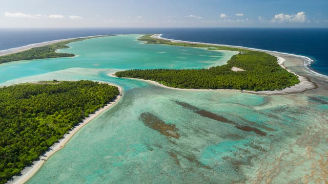Luftaufnahme einer tropischen Insel mit üppigem, grünem Wald, umgeben von türkisfarbenem Wasser. Die Küstenlinie ist von weißen Sandstränden gesäumt, und das Meer wechselt von hellen zu dunkleren Blautönen. Im Hintergrund erstreckt sich der Ozean bis zum Horizont unter einem klaren, blauen Himmel.