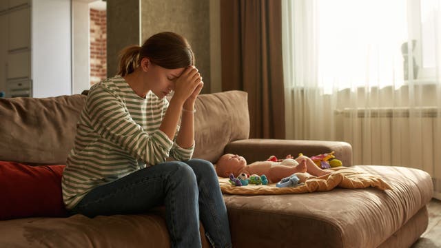 Eine Mutter sitzt auf dem Sofa neben ihrem Baby. Sie wirkt überfordert, besorgt und verzweifelt.
